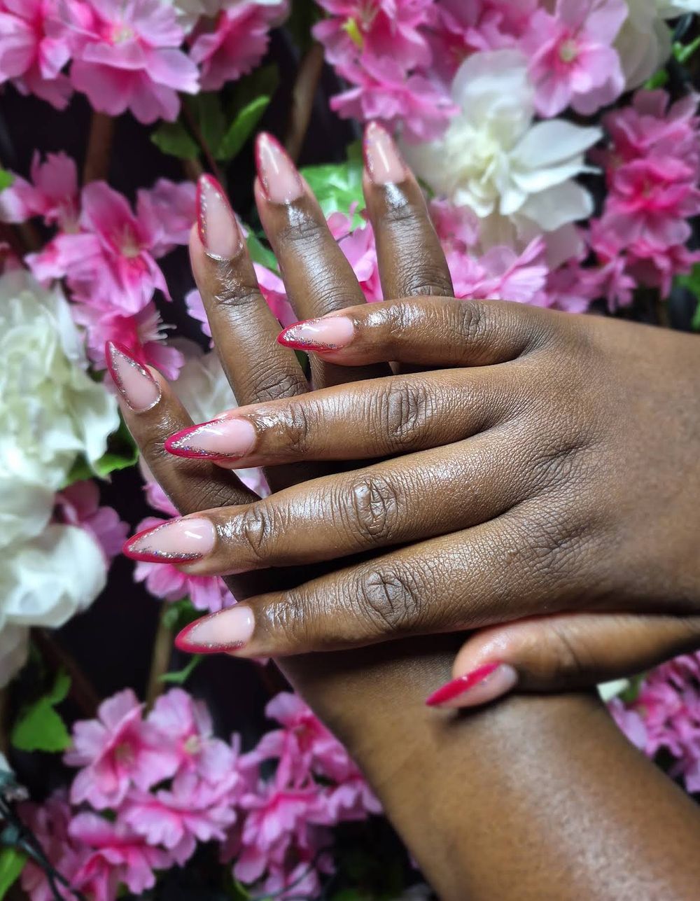 Nude almond nails with berry-red edges and silver glitter, held against pink blossoms